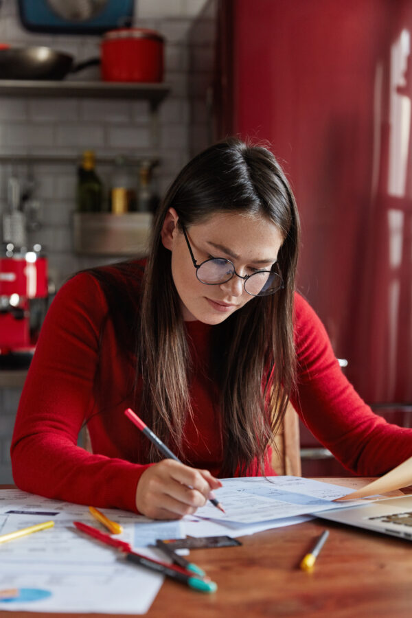 Busy female enterpriser thinks about target or planning company, makes statistics or analytic research, sits at desktop, kitchen interior in background, concentrated on work, wears round spectacles
